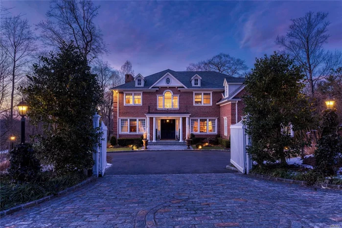 Tucked away behind white gates on a magical and secluded property, this 2007 brick Georgian Colonial offers you an exceptional opportunity to own a magnificent and pristine six-bedroom, five-and-a-half-bath home in an enchanting setting and an ideal location. With all the grace and charm of a “Golden Age” estate, this timeless beauty offers the comforts and amenities so desirable today. All this is off a winding wooded avenue in Roslyn Harbor, a small residential village overlooking the waters of Hempstead Harbor.  Originally a farming community, it was developed by wealthy New Yorkers into their summer estates.  Two of those properties remain today, the modern Engineer’s Country Club and the William Cullen Bryant Preserve with the Nassau County Museum of Art.  Located just north of Roslyn Village, the area is convenient to shopping (including the Americana at Manhasset and the Miracle Mile), fine dining, golf, boating, fabulous parks, railway station, and major highways, putting it within about 45 minutes from Manhattan. 
As you enter the courtyard of this beguiling residence, you may be intrigued by the sound of rushing water just out of sight. Pursuing your curiosity, you will find a waterfall cascading down several feet then tumbling along a stony creek bed to a wooded koi pond. There, you can enjoy a serene and quiet sitting area accessed from an enchanting stone bridal staircase. Perfectly built to transport you to a forest brook, these man-made wonders are just part of an acre of superbly designed landscaping with specimen trees and shrubs and a sweeping manicured lawn, all overlooked by an expansive raised stone patio.
The house is equally impressive with its coined brick façade, slate-like architectural roof, and welcoming wide portico. Double doors open beneath a towering Palladian clerestory to an enthralling two-story center-hall foyer with graceful staircase and heated inlaid-marble floors. Coffered ceiling, fielded-panel walls, and stunning classic moldings are indicative of the quality and character of the entire home. Flanked by the formal living and dining rooms, the foyer opens through a barrel-ceiling alcove, with powder room and closet, into a hallway accessing the kitchen and great room. This sunny and inviting gathering place enjoys a wood-burning fireplace, oak floors, coffered ceiling, three sets of French doors to the rear patio and large doorways to the living room and kitchen. Elegant custom cabinetry with crown molding and marble countertop creates a bright and airy feeling to this delightful workspace, boasting a large center island with corbeled seating area and prep sink. High-end appliances include a Bosch dishwasher and Viking refrigerator, microwave, convection oven, and gas range with six burners, two ovens, and pot filler. The cheerful breakfast area enjoys a custom built-in breakfront and French doors to the patio. Off the kitchen are a butler’s pantry, a walk-in pantry, laundry room, backstairs, maid’s quarters, study, access to the garage, and side entrance.
In its own wing of the second floor, the regal and aethereal master suite is a dream come true. With chandeliers hanging from high tray ceilings, and a cozy wood burning fireplace, the master bedroom and sitting room are joined by a coffered barrel-ceiling archway. A hall has marble floors extending into two walk-in closets and a luxurious master bath. This features radiant heated floors, a whirlpool tub, two console vanities, and a shower. Two additional bedrooms share a Jack-and-Jill bath while two more enjoy en suite baths along with a spacious bonus/play room with vaulted ceiling and dormers. A wine connoisseur’s entertainment paradise, the full finished basement boasts an immense entertainment space with “tin” ceiling, hand-painted vintner’s-sign mural, three sets of tall French doors opening to a patio overlooking the babbling brook, and an immense wine cellar with arched, planked entrance doors, stone arches, tile floors, and brick walls and ceiling, that can hold over 2,000 bottles of wine. Paying tribute to Long Island’s glorious past while offering an almost storybook setting of charm and romance, this unique home is truly qualified to be your forever-after home.